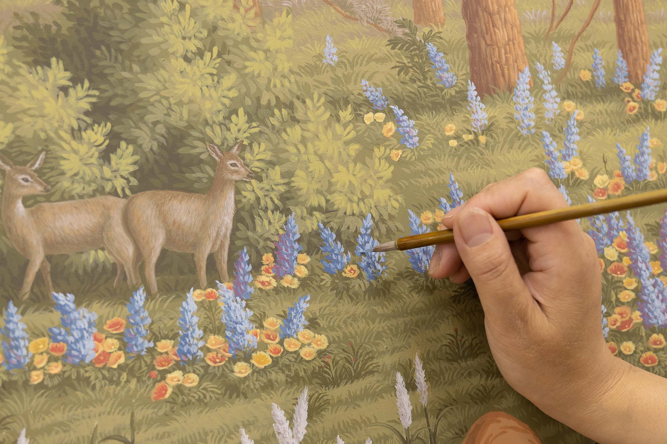 A hand holds a paintbrush, adding blue flowers to a detailed painting of a forest scene with two deer and colorful flowers among trees and foliage.