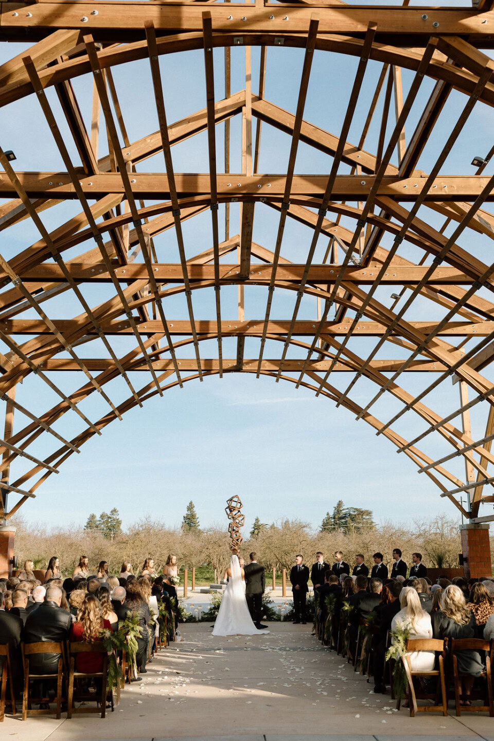 A bride and groom stand at the altar under a large wooden pavilion, surrounded by guests seated on either side. The outdoor wedding scene is bright and open, with trees and a blue sky in the background.