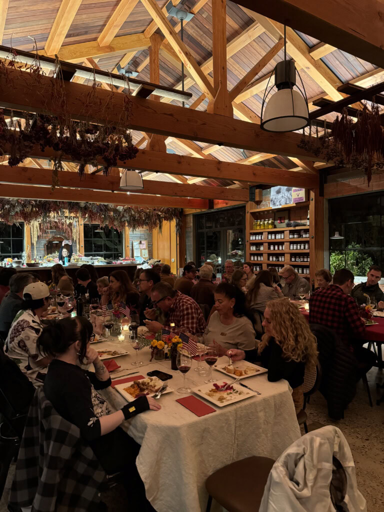A group of people dine together at long tables in a warmly-lit restaurant with wooden beams, dried herbs hanging from the ceiling, and shelves lined with jars in the background.
