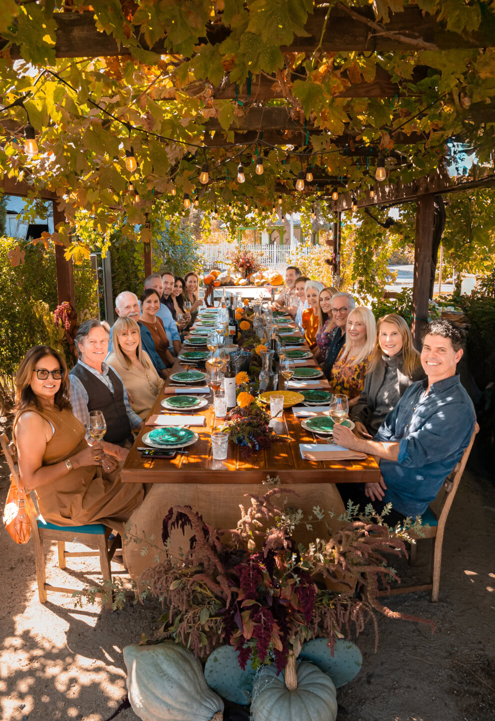 A group of people sit around a long outdoor dining table under a pergola with hanging lights and green vines, enjoying a festive meal on a sunny day. The table is set with plates, glasses, and autumn-themed decorations.