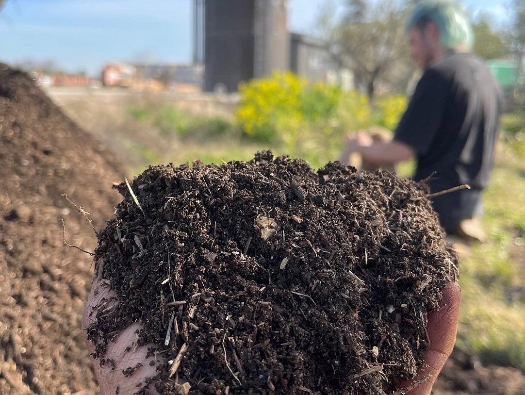 A close-up of a hand holding rich, dark soil outdoors, with blurred greenery and a person with light hair kneeling in the background on a sunny day.