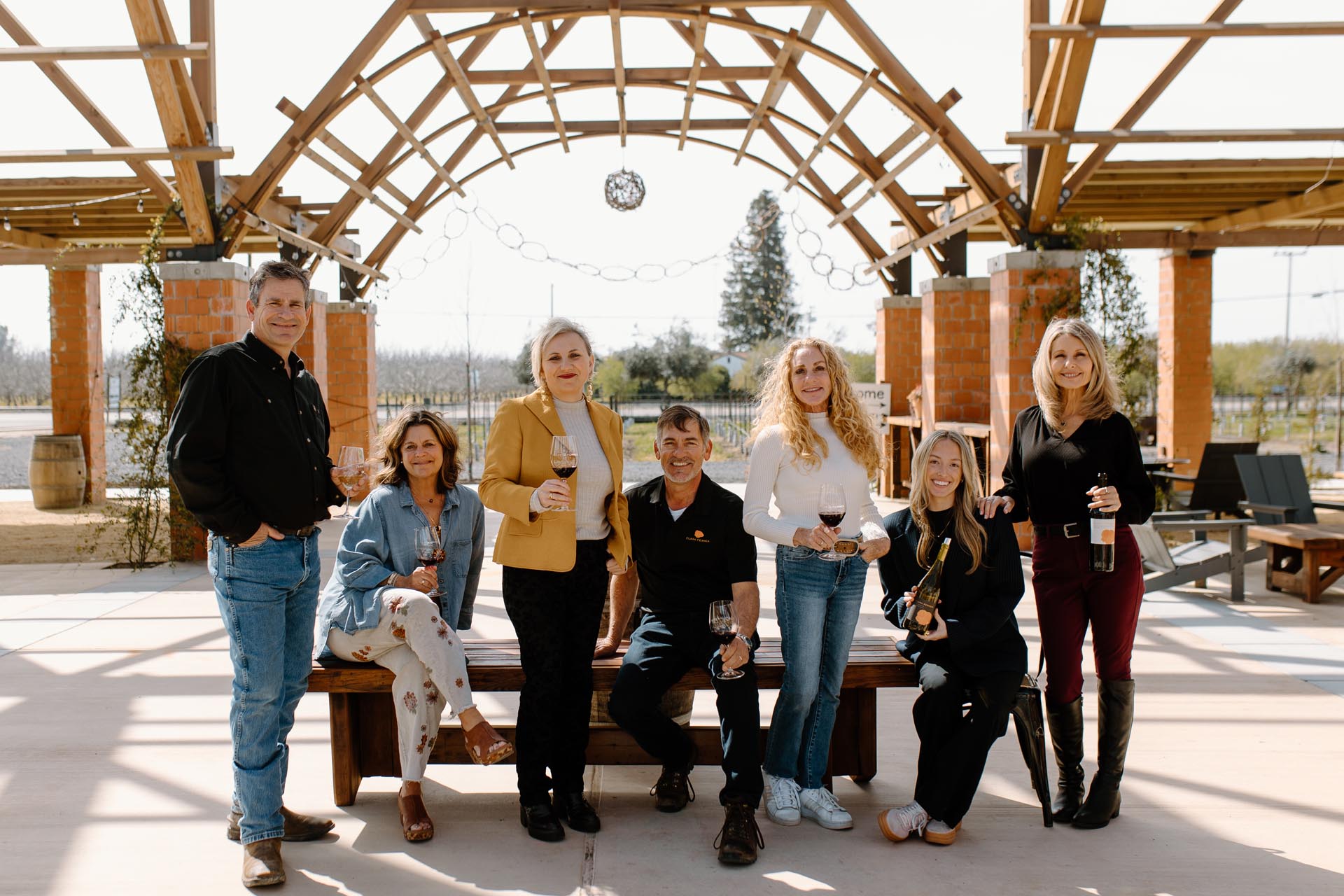 Seven adults stand and sit under a wooden pergola, holding glasses of red wine and smiling at the camera; it's a sunny outdoor setting with trees in the background.