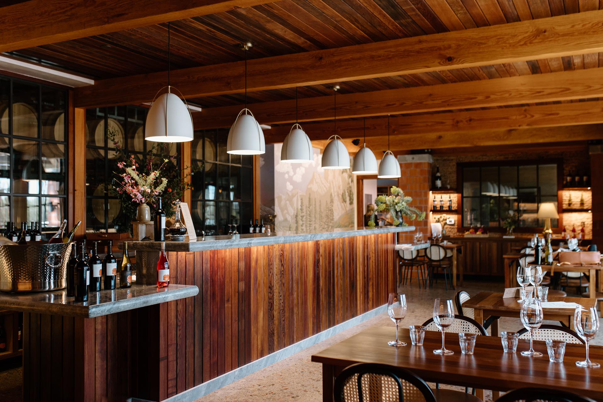 A stylish tasting room interior with a long wooden bar, modern pendant lights, wine bottles, and neatly set dining tables with glasses. The room features wood beams and large windows, creating a warm and inviting atmosphere.