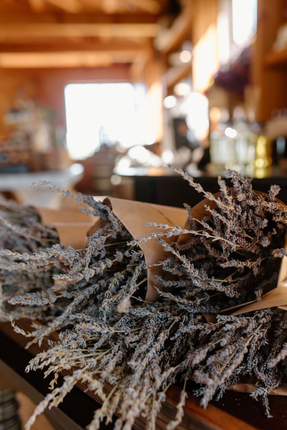 A close-up of dried lavender bouquets wrapped in brown paper, displayed on a wooden table in a warmly lit shop with blurred shelves and bottles in the background.