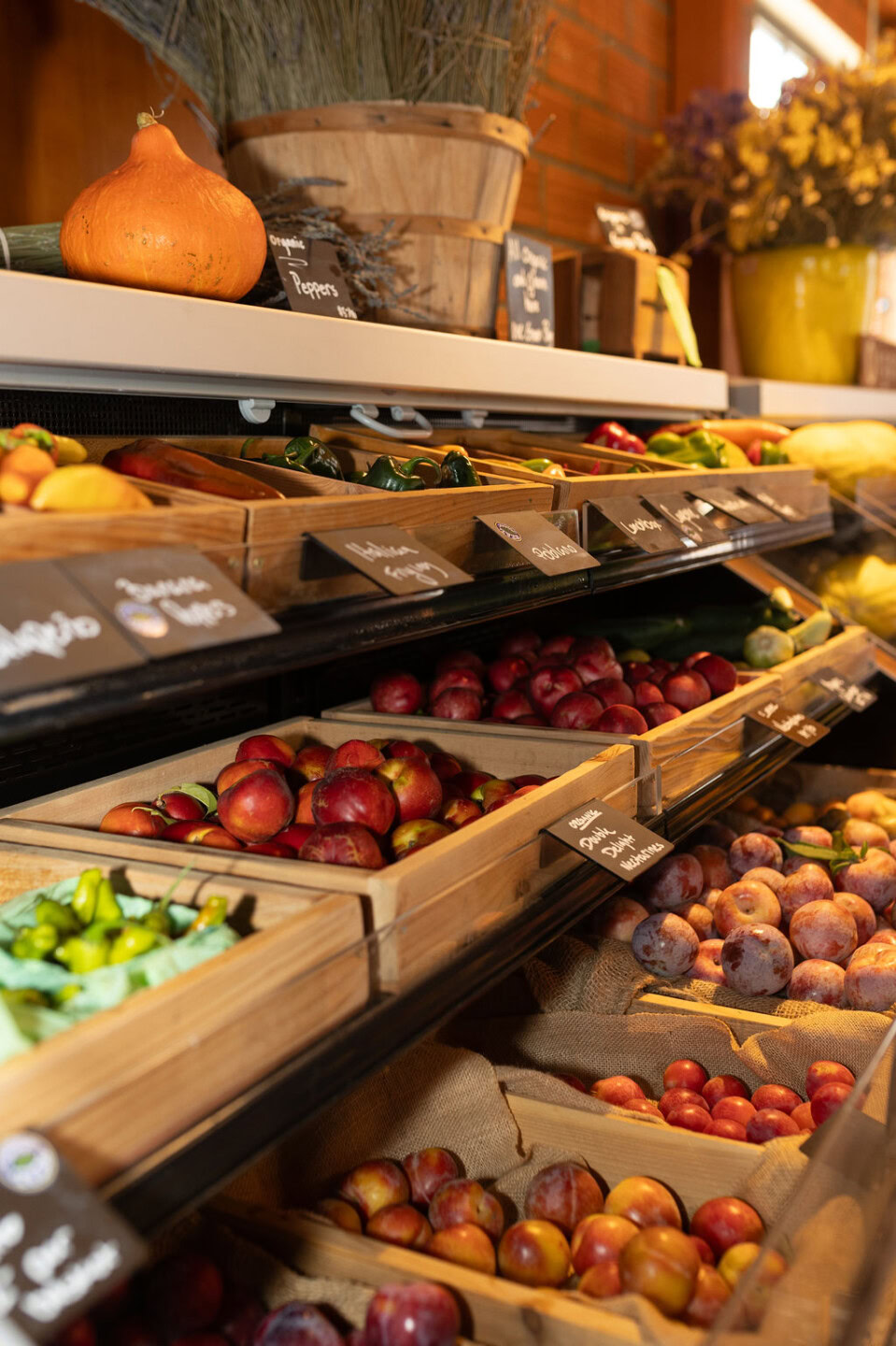 Wooden crates filled with fresh produce, including apples, plums, and cucumbers, are displayed on shelves in a market. Other fruits and vegetables are visible with chalkboard labels, and dried lavender sits above.