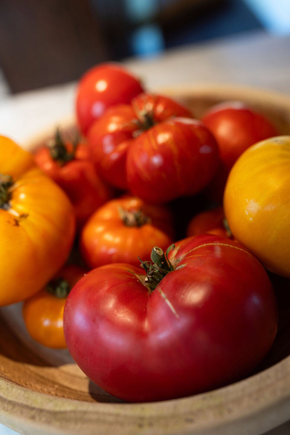 A wooden bowl filled with assorted heirloom tomatoes in shades of red, yellow, and orange, sitting on a light surface with a soft, blurred background.