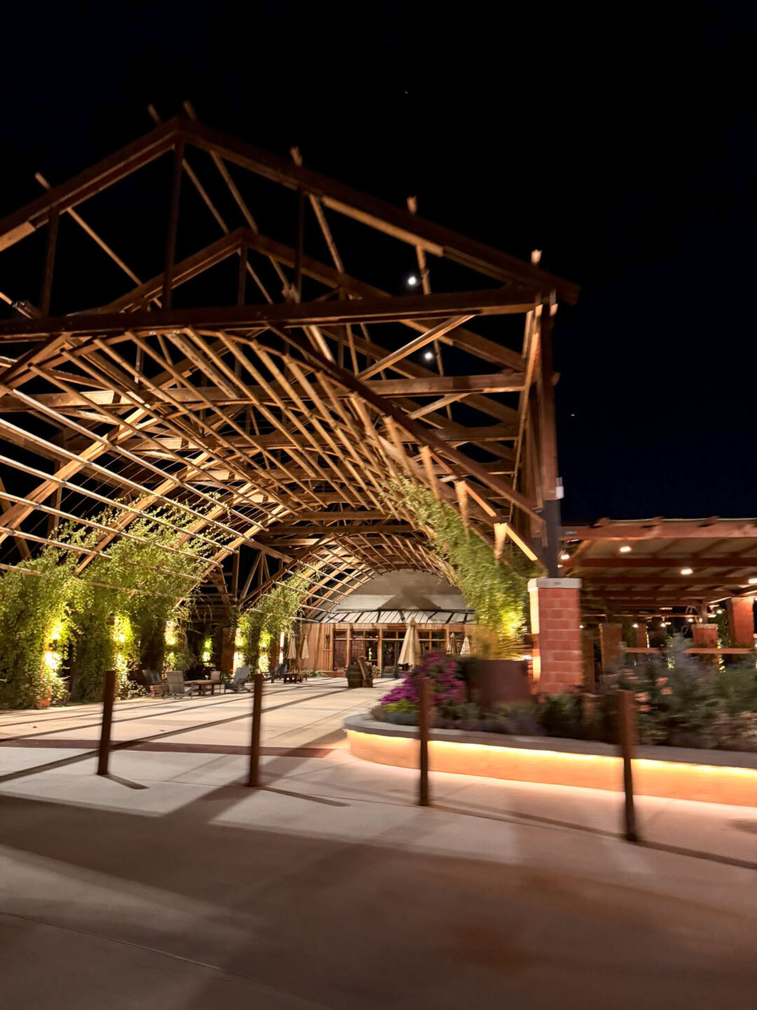 A large, open wooden pavilion structure with exposed beams, greenery, and warm night lighting, creating an inviting atmosphere along a pathway under the dark night sky.