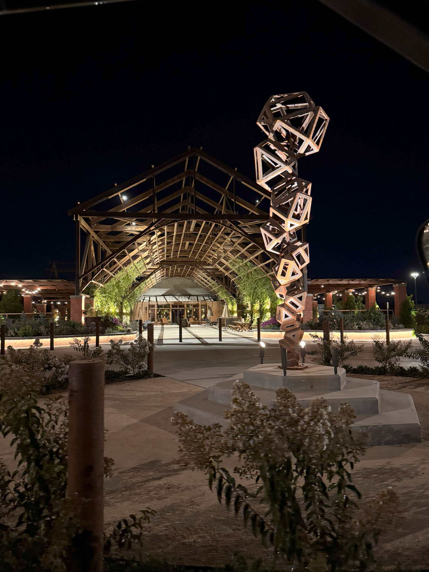 A wooden geometric sculpture stands in front of a modern building with a large, arched wooden entrance, illuminated at night with greenery and lights along the pathway.