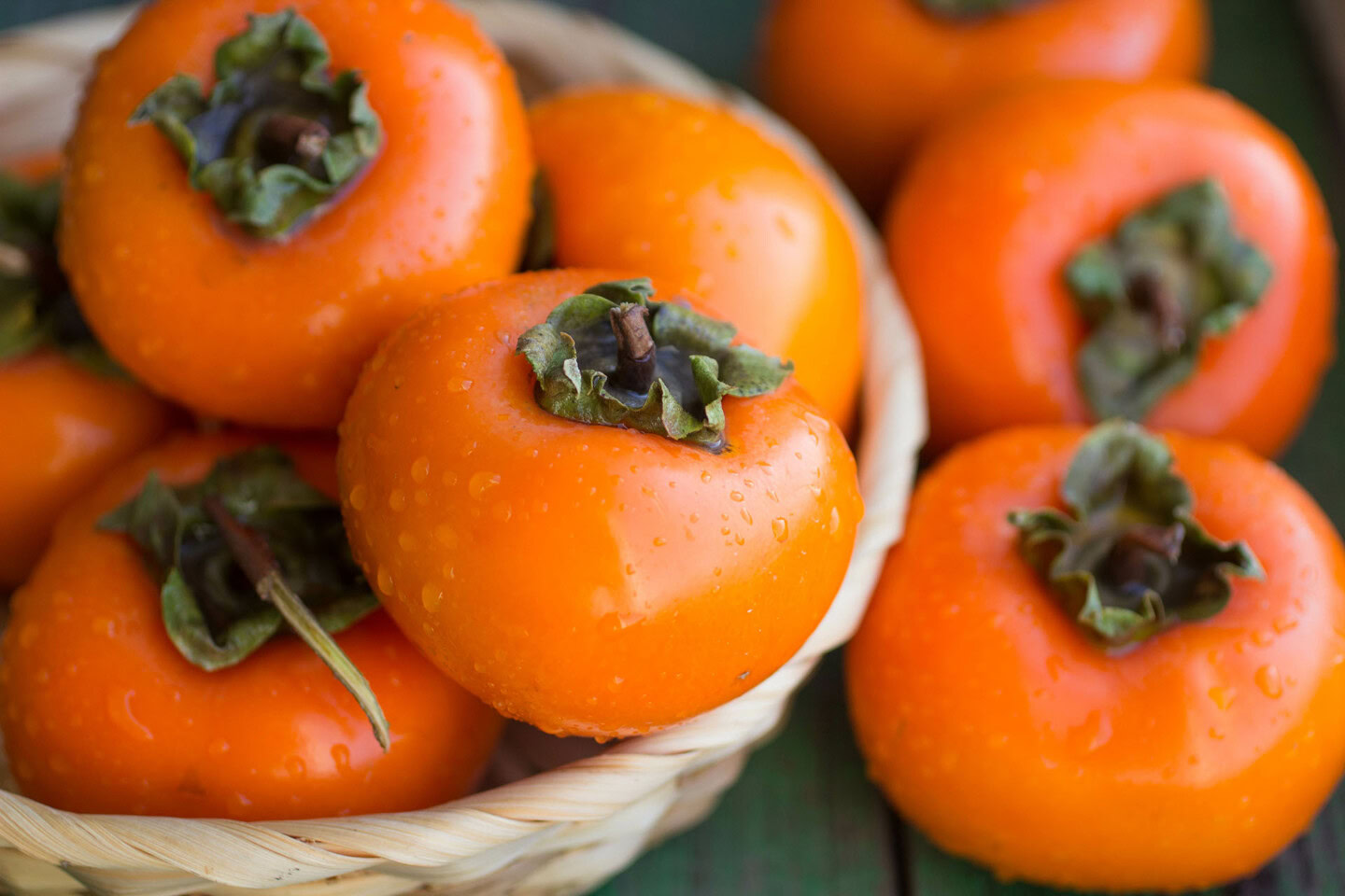 A close-up of ripe orange persimmons with green leafy tops, some inside a woven basket and others placed beside it, with droplets of water visible on their smooth skins.