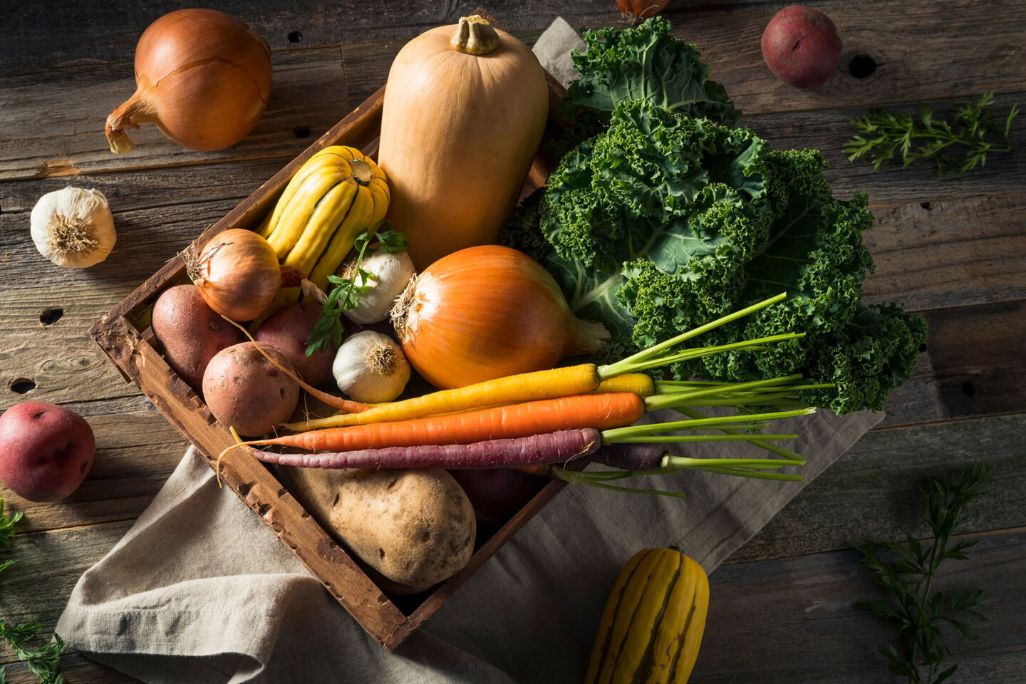 A wooden box filled with assorted fresh vegetables, including onions, garlic, potatoes, carrots, kale, butternut squash, and delicata squash, sits on a rustic wooden table.