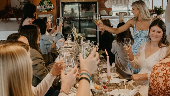 A group of women sit around a long table at a restaurant, raising glasses in a toast, smiling and enjoying a celebratory meal together. The table is decorated with flowers and dishes.