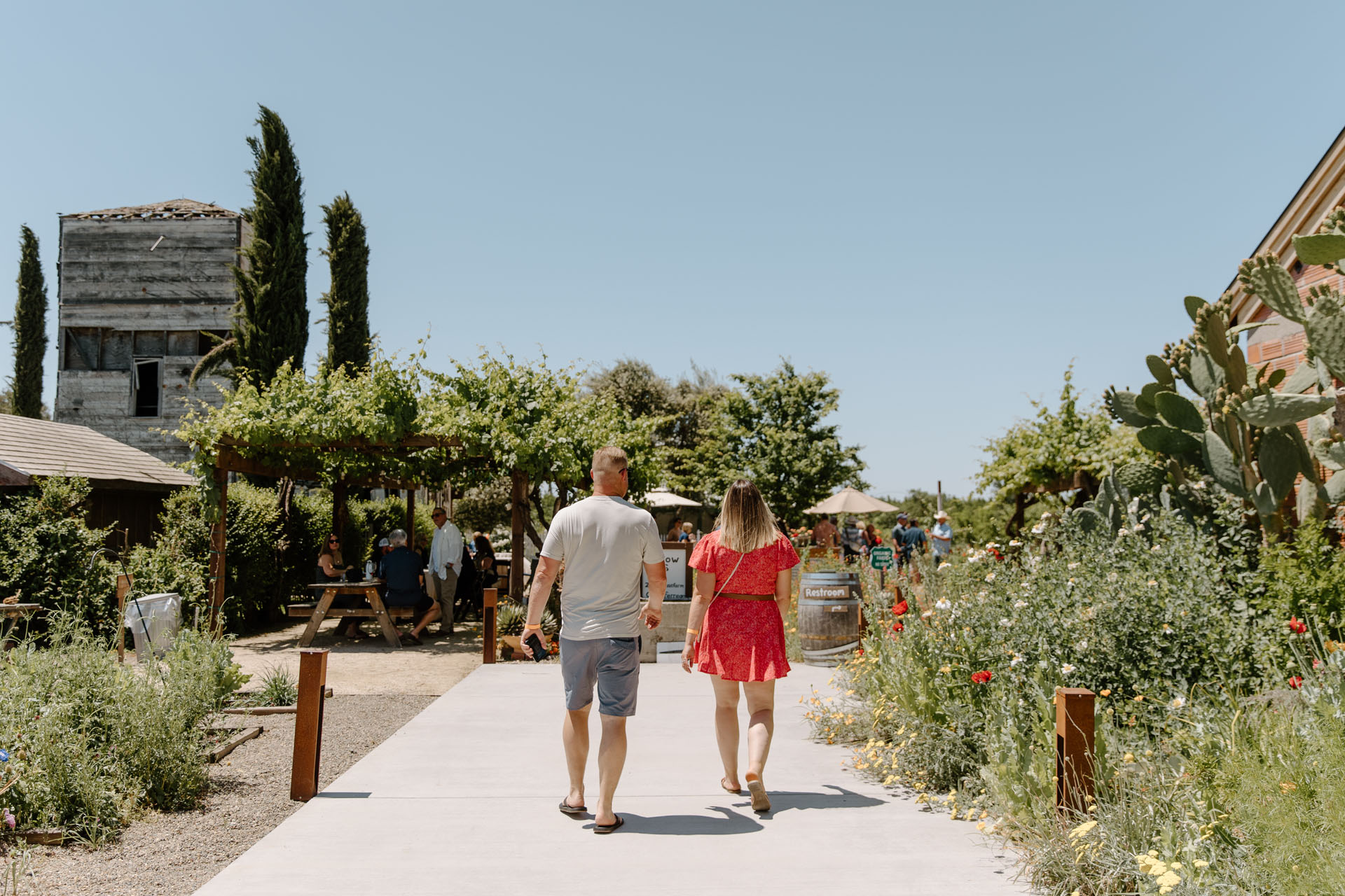 A man and woman walk hand in hand down a gravel path lined with greenery and flowers at an outdoor venue on a sunny day, with other people sitting at picnic tables in the background.