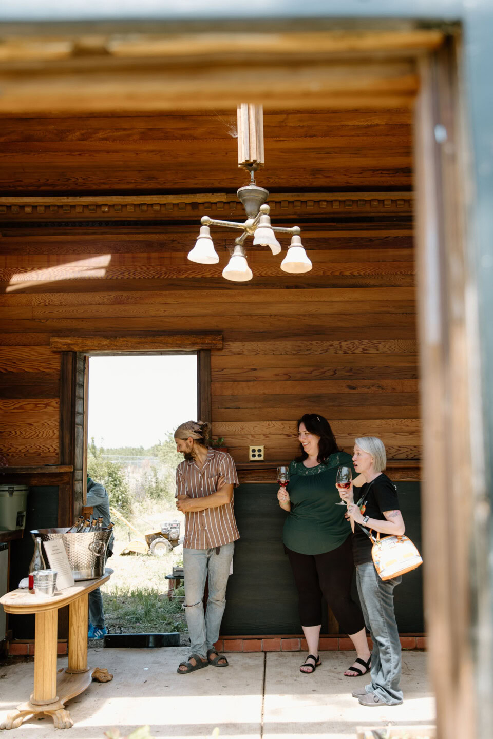 Three people stand inside a wooden room near an open doorway; two women hold drinks and chat, while a man leans against the wall, looking down. A small table with items is visible on the left.