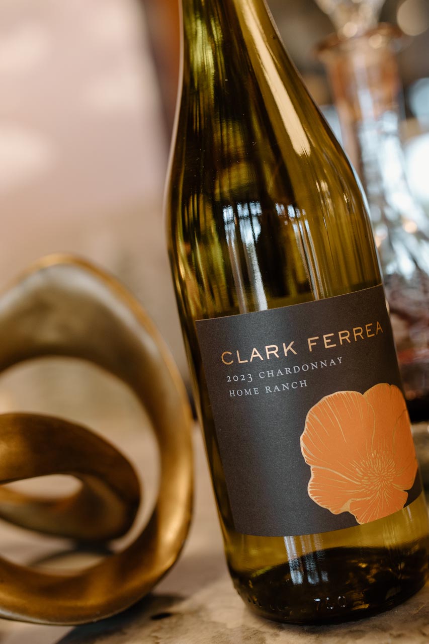 A close-up of a Clark Ferrea 2023 Chardonnay wine bottle with a dark label featuring an orange poppy illustration. The bottle is on a textured surface next to a modern gold sculpture.