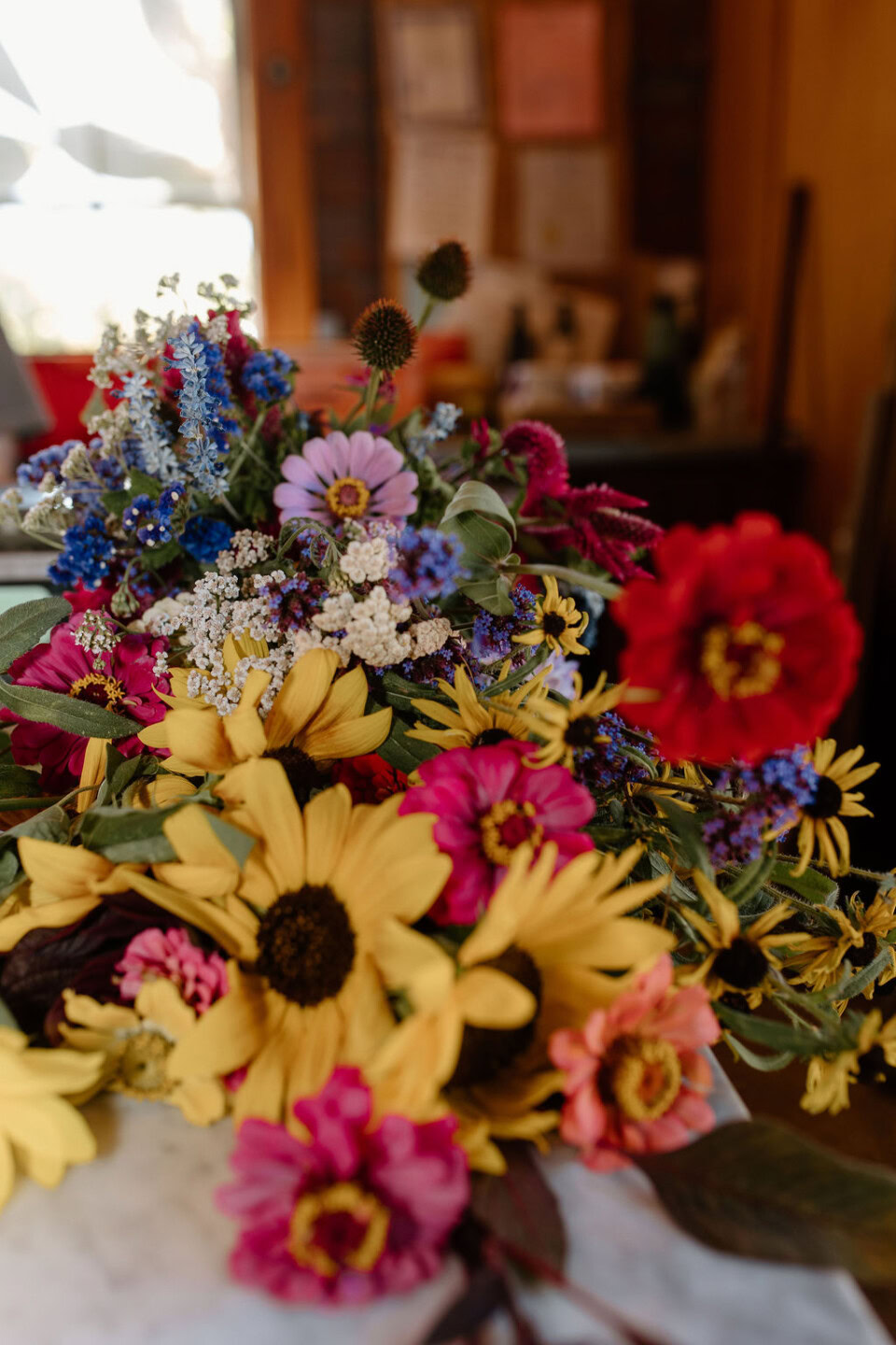 A vibrant bouquet of assorted flowers, including yellow sunflowers, pink zinnias, red blossoms, and small blue and white flowers, lies on a white surface in a warmly lit room with a blurred background.