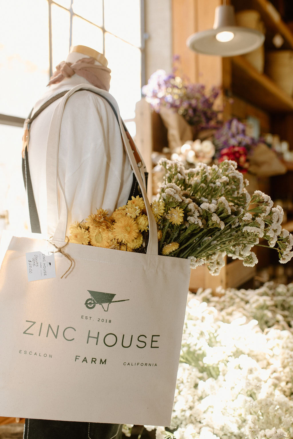 A mannequin dressed in a scarf and apron holds a tote bag filled with yellow and white flowers. The bag reads "Zinc House Farm, Escalon, California." The background features shelves with more flowers and warm sunlight.