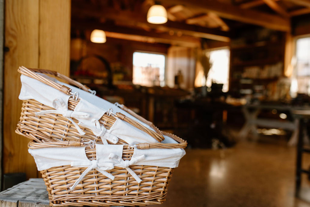 Two empty wicker baskets with white cloth linings and bows are stacked on a wooden surface inside a warmly lit rustic store or market with wooden beams and shelves in the background.