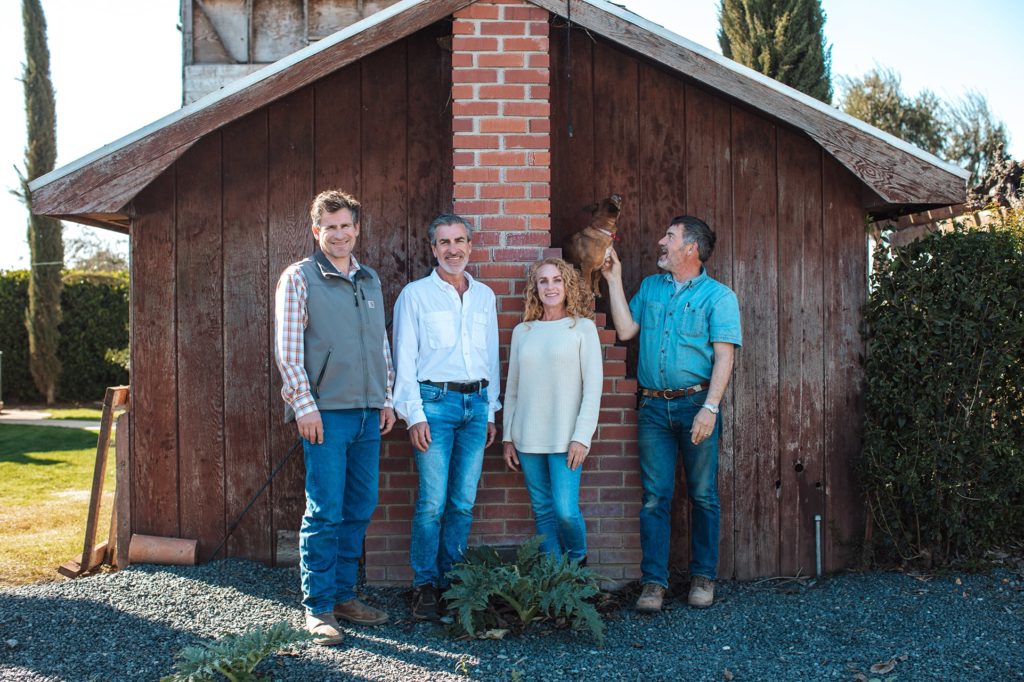 Four adults stand in front of a wooden shed with a brick chimney. One man points to a chicken perched on the chimney. They are smiling and dressed in casual clothes. Bushes and trees are visible in the background.