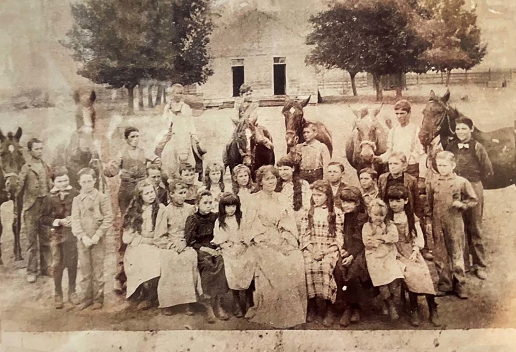 A sepia-toned vintage photo of a large group of children and a few adults posing outdoors in front of a building, with several horses in the background and trees nearby. The children wear old-fashioned clothing.