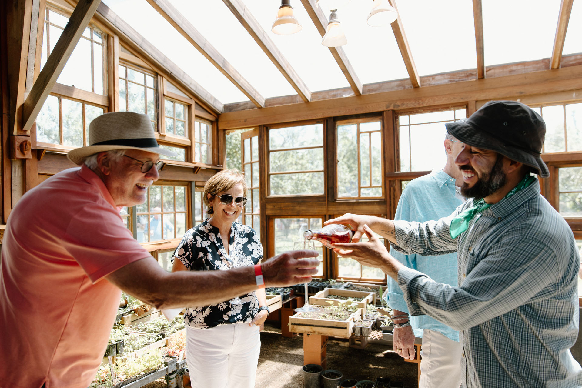 A smiling man in a hat pours a drink for an older man in a sunhat inside a bright greenhouse, while two other people, a woman and another man, watch and smile. Sunlight streams through the glass roof and windows.