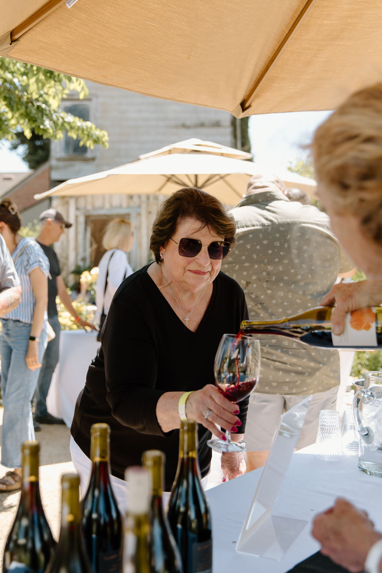 An older woman wearing sunglasses smiles as someone pours red wine into her glass at an outdoor event, with wine bottles on a table and people mingling under umbrellas in the background.