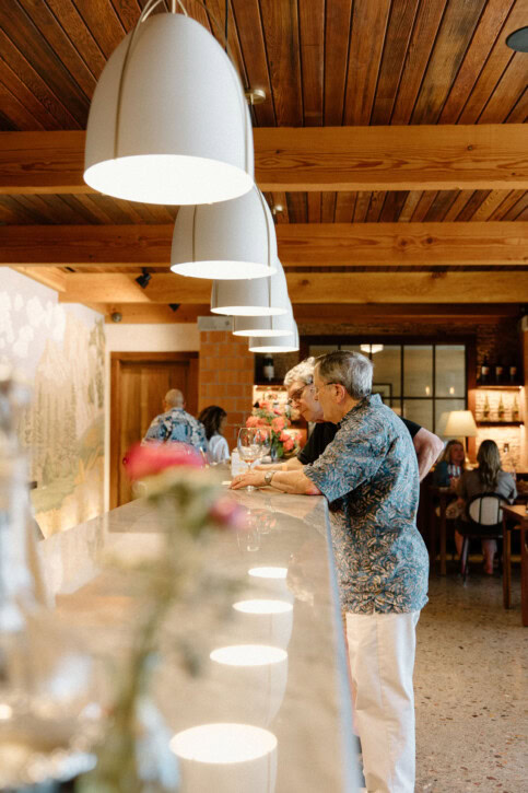 People stand at a modern bar with a wooden ceiling and pendant lights. A blurred flower is in the foreground, while others socialize in a warmly lit, inviting atmosphere in the background.