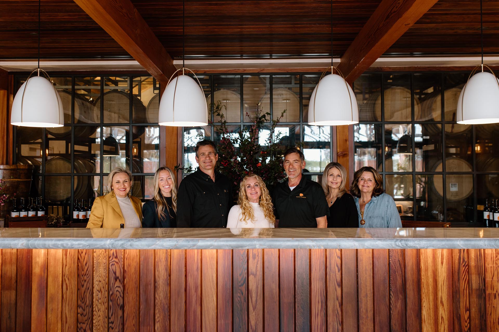 Seven adults stand smiling behind a wooden counter in a winery tasting room, with wine bottles and large barrels visible in the background and three white pendant lights hanging above.